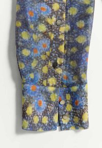 Manche d'une chemise multicolore avec un motif floral abstrait jaune, bleu et orange et des boutons-pression argentés sur fond blanc.