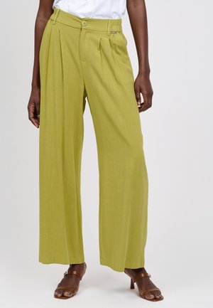 mellemhøje, vidtbenede grønne bukser med forreste læg, båret med hvid top og brune sandaler med hæl på person med mørk hud, der står