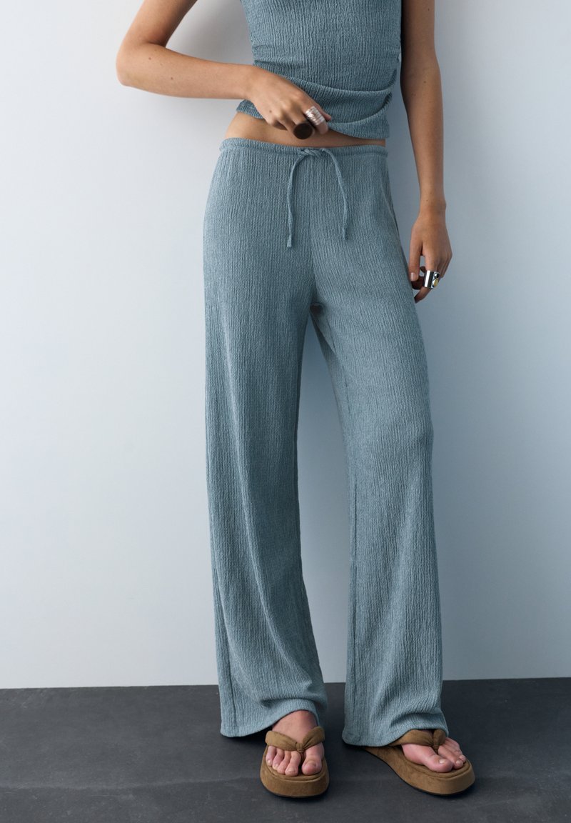 Femme portant un pantalon ample bleu texturé avec taille à cordon, un haut sans manches assorti, de grandes bagues, et des sandales compensées marron.