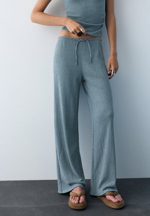 Kvinde iført løstsiddende blå strukturerede bukser med snøre i taljen, matchende ærmeløs top, store ringe og brune platformsandaler.