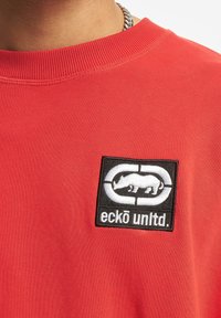 Punainen puuvillaneule, jossa on musta brodeerattu logo, jossa on valkoinen sarvikuono-siluetti ja teksti "eckō unlt.d." alapuolella.