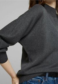 Pull en tricot gris avec un col montant, une coupe ample et des poignets côtelés. Porté avec un jean en denim bleu, montrant une ceinture noire.