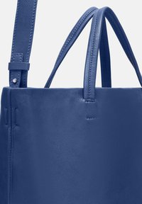 Sac fourre-tout en cuir bleu avec une texture lisse. Il possède deux longues poignées et un design minimalist avec des coutures visibles et une fermeture sécurisée.