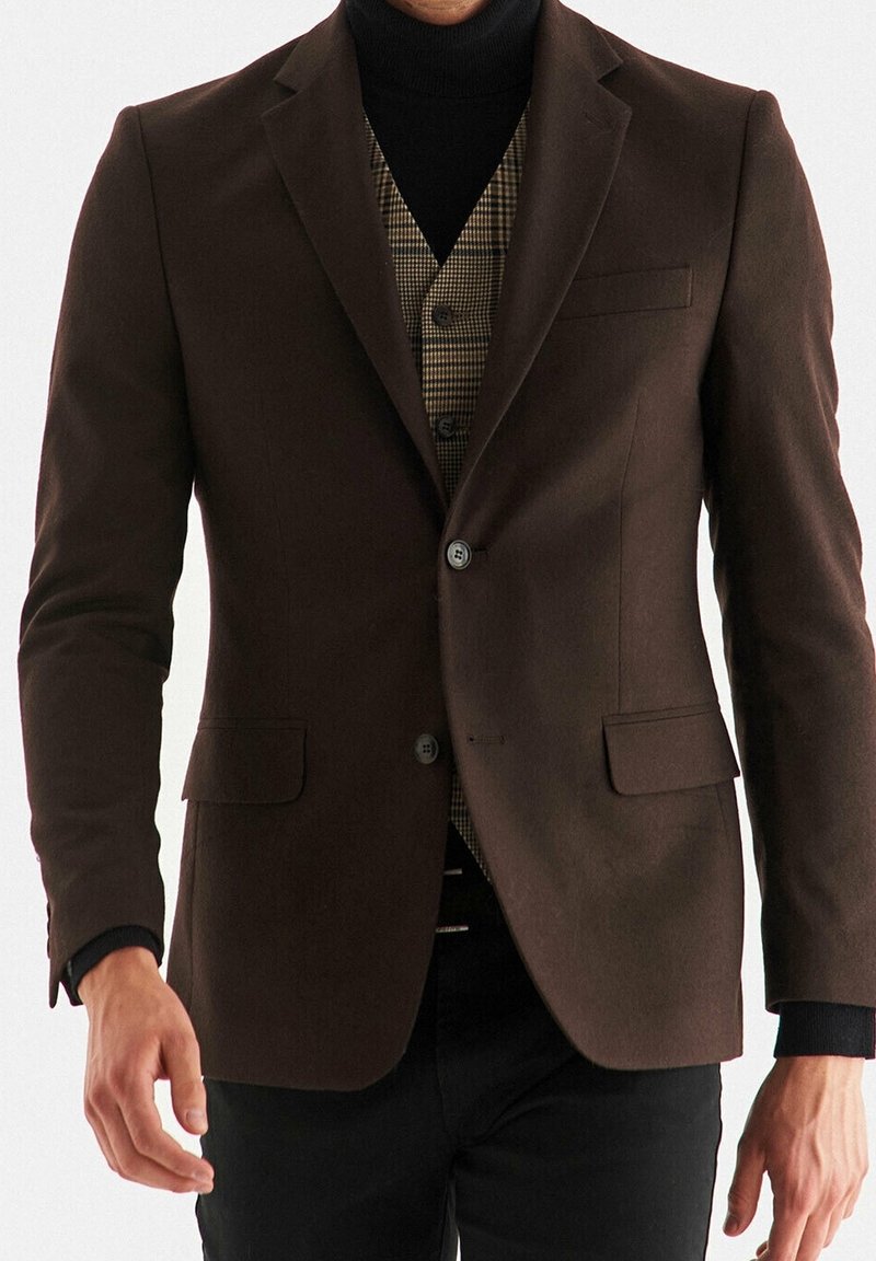 Blazer marrone su misura con rever a punta, chiusura con un bottone e due tasche anteriori, indossato sopra un gilet a quadri.