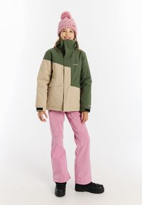 Grün-beige farblich abgesetzte Jacke mit hohem Kragen, kombiniert mit pinken Hosen und schwarzen Schuhen. Auffälliger Pom-Pom-Hut in hellpink.