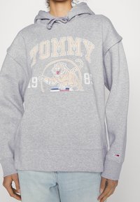 Harmaa huppari, joka on valmistettu pehmeästä kankaasta ja jossa on kohopainatus tiikeristä, "TOMMY"-teksti sekä pieni logo-merkki hihoissa.