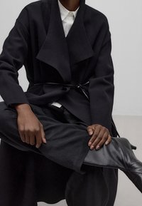 Musta ylipitkä takki, jossa on lovikaulus ja ohut solmittava vyö. Sileä pinta, yhdistetty istuviin mustiin housuihin ja korkeisiin mustiin saappaisiin.