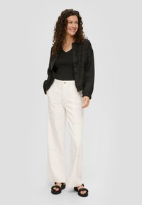 Czarna kurtka ze zapięciem na guziki noszona na czarnej, prążkowanej bluzce, w połączeniu z kremowymi spodniami o szerokich nogawkach z detalami szwów i czarnymi sandałami na platformie.