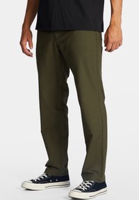 Homme portant un pantalon droit vert olive avec des baskets décontractées noires et blanches sur fond blanc.
