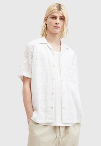 Hvid kortærmet skjorte med et struktureret mønster, knaplukning i front med træknapper og en afslappet krave. Bæres over en hvid tanktop.