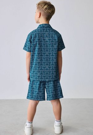 Junge steht mit dem Rücken zur Kamera, trägt ein passendes blau gemustertes kurzärmeliges Hemd und eine kurze Hose sowie weiße Turnschuhe und Socken in einem schlichten Raum.