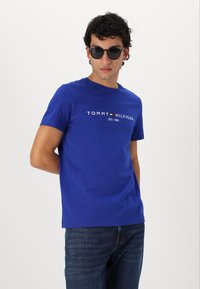 Blaues Baumwoll-T-Shirt mit Rundhalsausschnitt, mit dem weißen "TOMMY HILFIGER"-Logo quer über die Brust und in normaler Passform. Getragen mit dunklen Jeans.