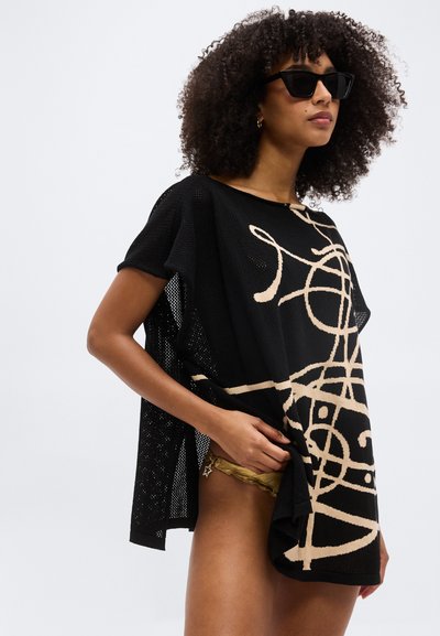 Γυναίκα με σγουρά μαλλιά που φοράει μαύρα γυαλιά ηλίου και φαρδιά μαύρη μπλούζα με μπεζ αφηρημένα σχέδια, κρατώντας το τελείωμα στη μία πλευρά.