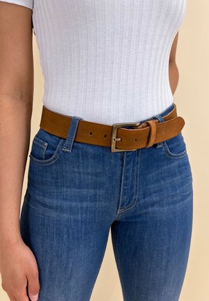 Personne portant un jean bleu et un haut blanc à manches courtes côtelé avec une ceinture en cuir marron attachée à la taille.