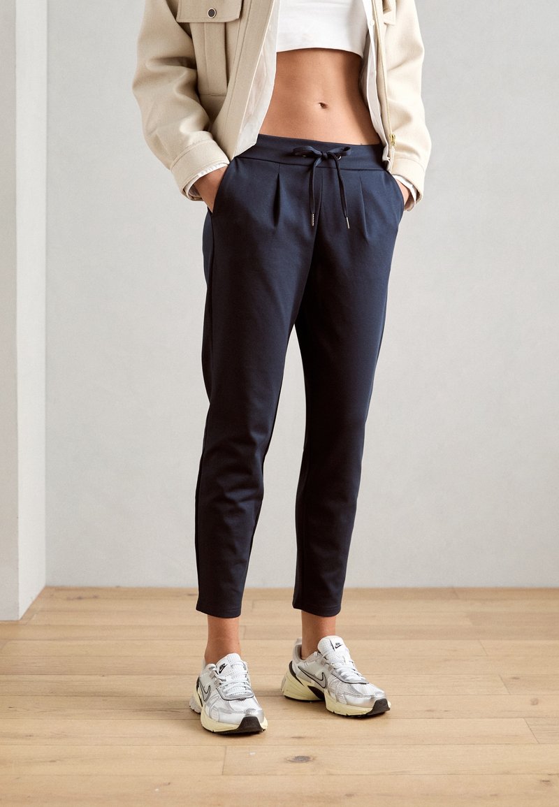 Pantalon de jogging bleu marine à taille cordon, coupe ajustée et plis, assorti à des baskets blanches. Le modèle porte un top court et une veste légère.