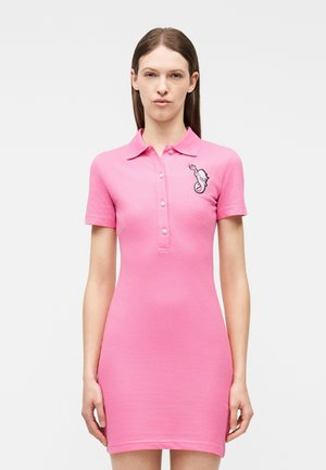 Kvinde iført en tætsiddende pink polokjole med korte ærmer, knaplukning og et søhesteemblem på venstre bryst.