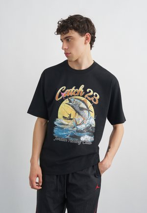 Young man in black shirt with large graphic of a jumping fish, fishing boat, and text "Catch 23 Jordan Fishing Club."