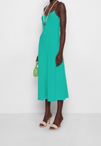 Femme portant une robe midi sans manches en teal, des sandales à talons beiges, tenant un petit sac à main vert et portant un long collier de perles.