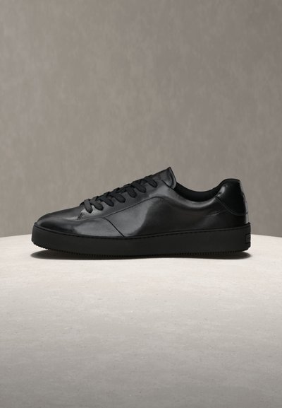 Μαύρο δερμάτινο χαμηλό αθλητικό παπούτσι με μαύρα κορδόνια και σόλα, παρουσιάζεται σε φωτεινή υφή επιφάνειας σε απλό γκρι φόντο.