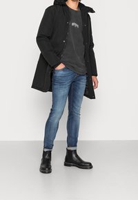 Černý prošívaný kabát s kapucí, šedé tričko s potiskem, modré džíny se slim střih, a černé Chelsea boty s texturovanou podrážkou a kontrastním prošíváním.