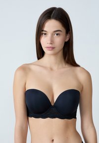 Reggiseno nero senza spalline con tessuto liscio opaco e orlo in pizzo sul fondo. Le coppe sono leggermente imbottite con design senza cuciture.