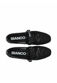 Zapatos Mary Jane de malla negra con punta de charol y correa, que presentan una forma cuadrada y suela plana, con un interior texturizado y marca blanca.