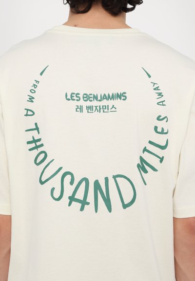 T-shirt crème avec du texte courbé en vert : "LES BENJAMINS" et "FROM A THOUSAND MILES AWAY", avec une texture douce et lisse.