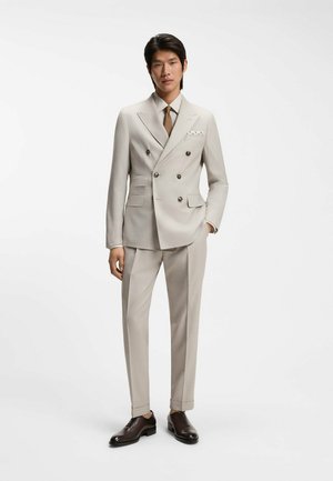 Uomo che indossa un completo doppiopetto beige con cravatta marrone, camicia a righe, fazzoletto da taschino e scarpe eleganti marrone scuro, in piedi su sfondo bianco.