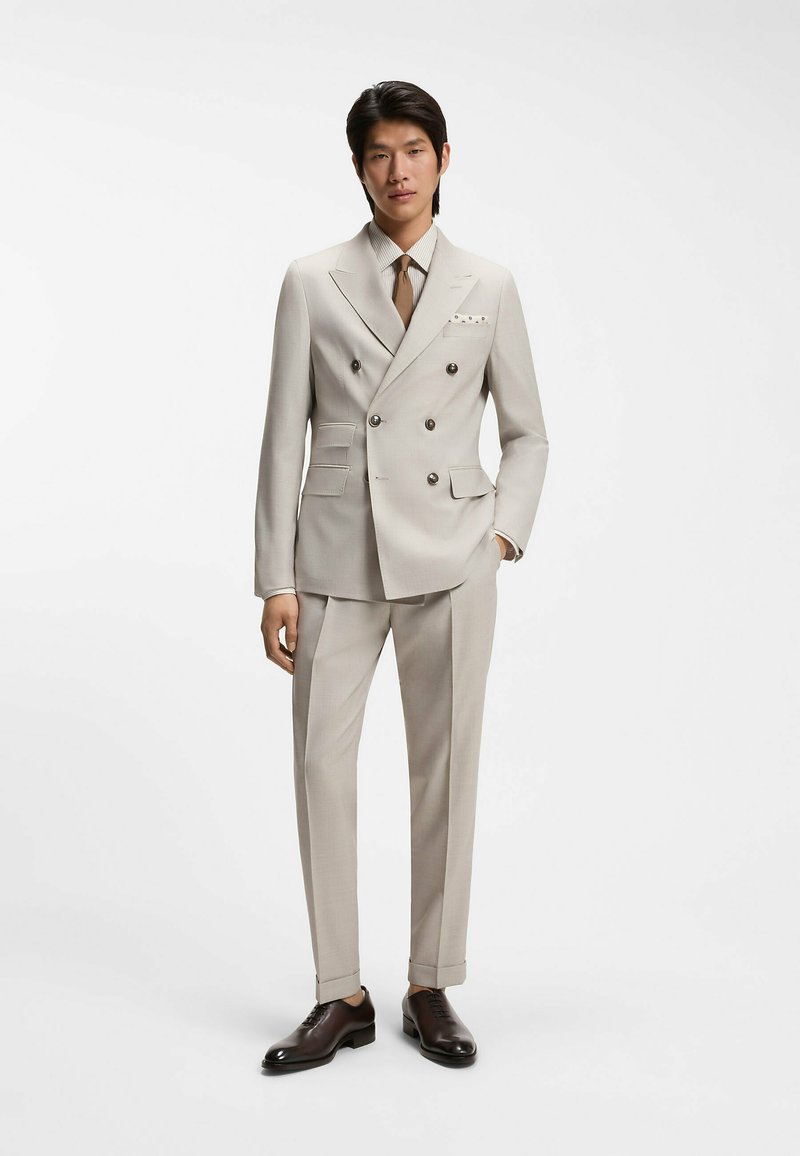 Homme portant un costume croisé beige avec une cravate marron, une chemise rayée, un mouchoir de poche et des chaussures habillées marron foncé, debout sur un fond blanc.