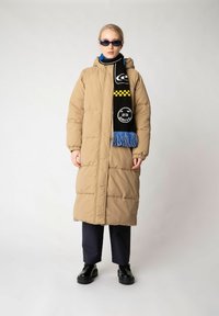 Beige, padded long coat with a hood, black and yellow patterned scarf, blue top underneath, and black pants, paired with black shoes.