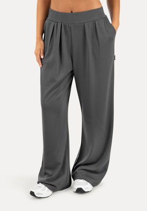 Pantalon ample gris foncé avec plis porté avec des baskets blanches, mains dans les poches, montré de la taille aux pieds sur un fond clair.