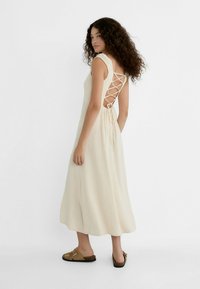 Beige linen dress with a fitted bodice and a lace-up back. Flat hemline and shoulder ruffle details. Worn with brown slip-on sandals.