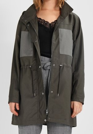 Chaqueta exterior de color verde oscuro con bolsillos grises, cintura con cordón y un alto cuello. Fabricada con un tejido ligero y con cierres de botones a presión.