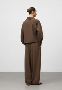 Giacca marrone oversize con colletto, abbinata a pantaloni marroni a gamba larga. Texture liscia, design minimale e dettaglio con bottone sul retro dei pantaloni.
