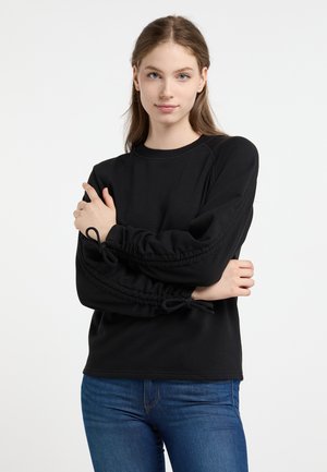 Felpa nera con maniche elastiche e arricciate legate con fiocchi. Presenta un classico collo rotondo e una texture morbida. Abbinata a jeans blu.