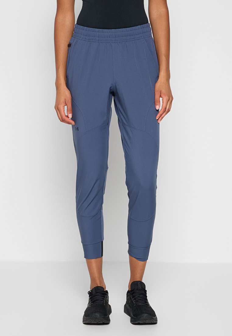 Personne portant un pantalon de sport bleu ajusté avec une taille élastique et des chaussures de sport noires, se tenant devant un fond clair.