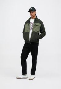 Chaqueta de forro polar en verde aceituna oscuro con cremallera frontal, bolsillos en contraste y textura suave. Combinada con una camiseta gráfica, pantalones negros y zapatillas blancas.