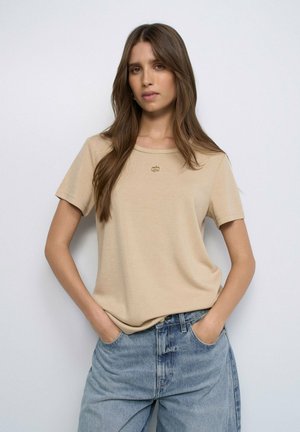Joven con cabello largo y castaño, viste una camisa beige de manga corta y vaqueros azules, de pie con las manos en los bolsillos sobre un fondo liso.