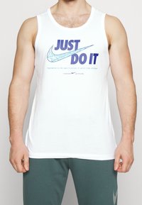 Man in een witte mouwloze Nike-shirt met "JUST DO IT" en een grafisch swoosh-ontwerp, gecombineerd met donkergroene trainingsbroek.