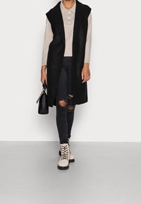 Schwarze lange Weste über einem beigen Pullover, kombiniert mit zerrissenen schwarzen Jeans und hellen Schnürstiefeln. Accessoire: kleine schwarze Handtasche.