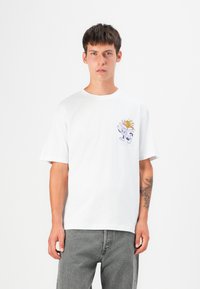 Weißes Baumwoll-T-Shirt mit Rundhalsausschnitt und kurzen Ärmeln, versehen mit einem bunten Grafikdruck auf der linken Brust, kombiniert mit grauen Jeans.