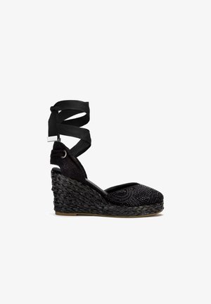 Espadrille noire à talon compensé avec une semelle en jute tissé, dotée d'un supérieur en tissu texturé orné de petites perles et d'une sangle de cheville en ruban croisées.