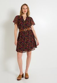 Robe noire avec des motifs géométriques colorés, manches courtes évasées, décolleté en V, taille cintrée avec une ceinture marron et jupe évasée.