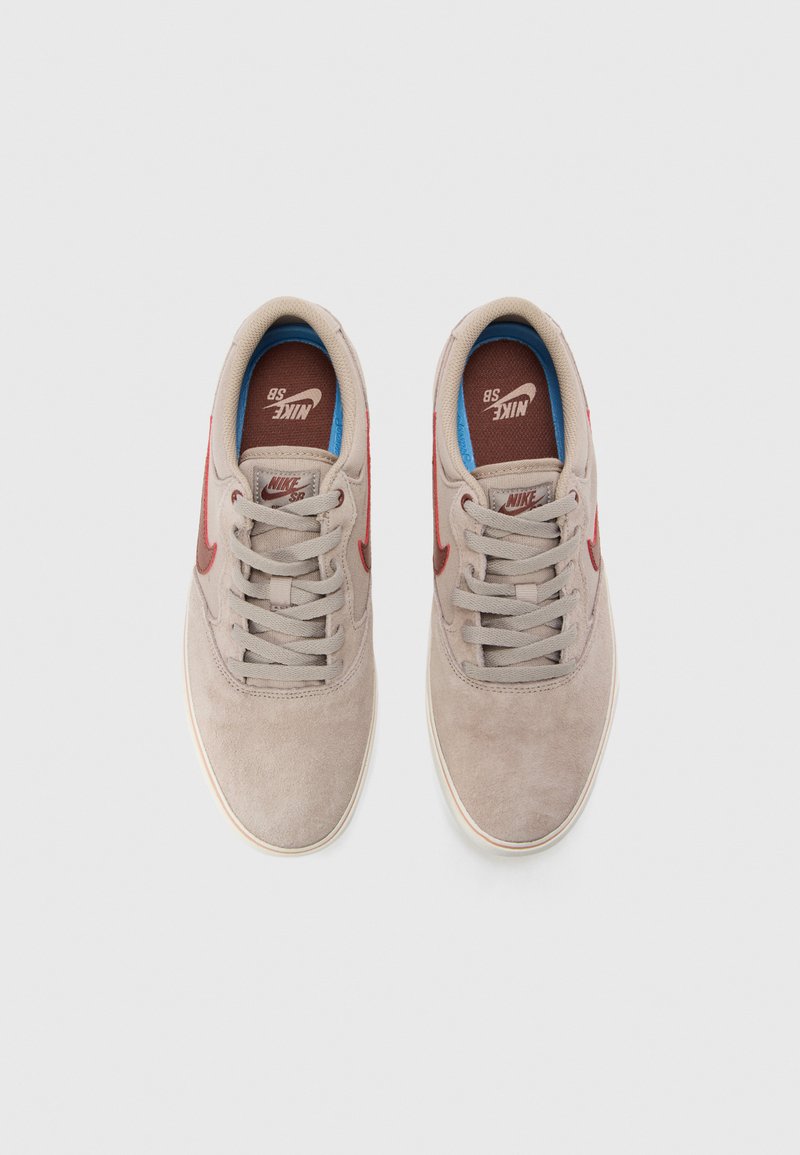 Beżowe zamszowe sneakersy z szarymi sznurówkami, czerwonym logo Nike, niebieską podszewką i białą gumową podeszwą. Czysty, minimalistyczny design.