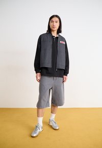 Gray zip-up vest with large pockets over a black shirt, paired with gray shorts and silver sneakers. Flat weave texture, athletic style.