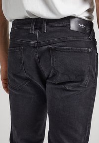 Svarta denimjeans med en figurnära passform, utrustade med fem fickor och en läderlogotyp på midjebandet. Diskret sömnadsdetalj synlig.