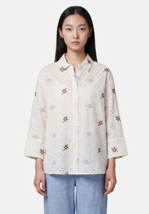 Femme portant une chemise blanche ample boutonnée avec des fleurs brodées marron et bleues et un pantalon large bleu clair.
