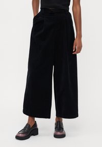 Sorte vide benbukser i kordfløjl med en tekstureret overflade, plisseret front, elastisk talje og sidelommer, parret med sorte loafers.