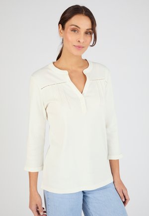 Maglia a maniche lunghe color crema con scollo henley, cuciture decorative e vestibilità leggermente morbida abbinata a jeans azzurri.