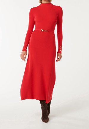 Kobieta ubrana w czerwoną, prążkowaną dzianinową midi sukienkę z długim rękawem, z cienkim paskiem w tym samym kolorze oraz brązowe zamszowe kozaki za kolano.